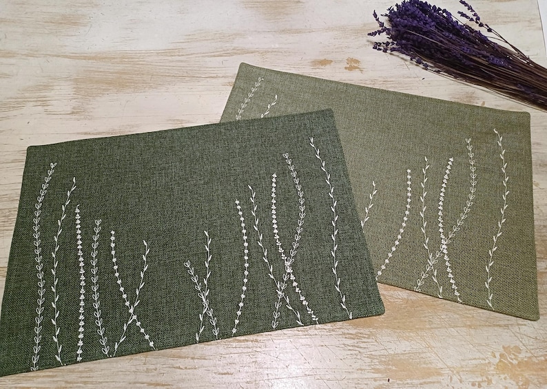 May include: Two green placemats with white embroidered floral designs. The placemats are made of a textured fabric.