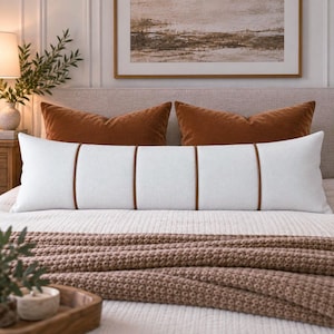 May include: A long, rectangular white lumbar pillow featuring a textured fabric surface and four vertical brown leather-like piping accents that divide the pillow into five distinct sections, positioned horizontally across a bed with bedding.