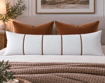 May include: A long, rectangular white lumbar pillow featuring a textured fabric surface and four vertical brown leather-like piping accents that divide the pillow into five distinct sections, positioned horizontally across a bed with bedding.