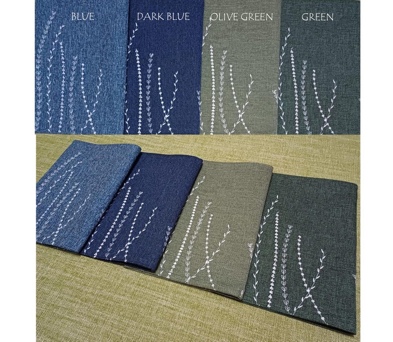 May include: Four placemats in blue, dark blue, olive green, and green with a white embroidered floral design. The placemats are laid out on a beige surface.