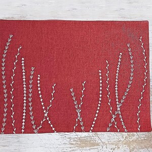 May include: A red placemat with a white embroidered design of stylized plants. The placemat is made of a textured fabric.