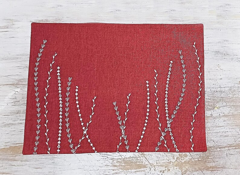 May include: A red placemat with a white embroidered design of stylized plants. The placemat is made of a textured fabric.
