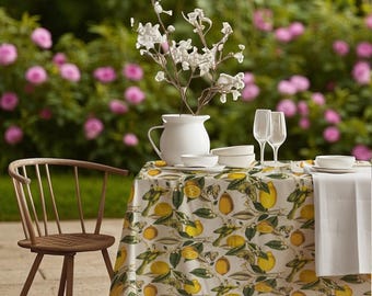 Puede incluir: Una mesa de comedor preparada para una comida al aire libre. La mesa está cubierta con un mantel con estampado de limones y decorada con vajilla blanca, copas de vino y una jarra blanca con tallos florales. Una silla de madera está al lado, con flores rosas de fondo.