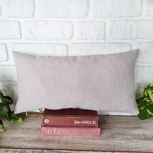 May include: A light gray linen throw pillow resting on top of two books. The top book is red and has the text "Sato + Kafka" on the spine. The bottom book is a dark red color.
