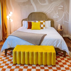 May include: A white bed with a brown headboard and a yellow tufted bench in front of it. The bed has a white blanket and a brown throw blanket. There are pillows on the bed in shades of brown, yellow, and white. The wall behind the bed is painted a light brown color and has a white abstract design on it.