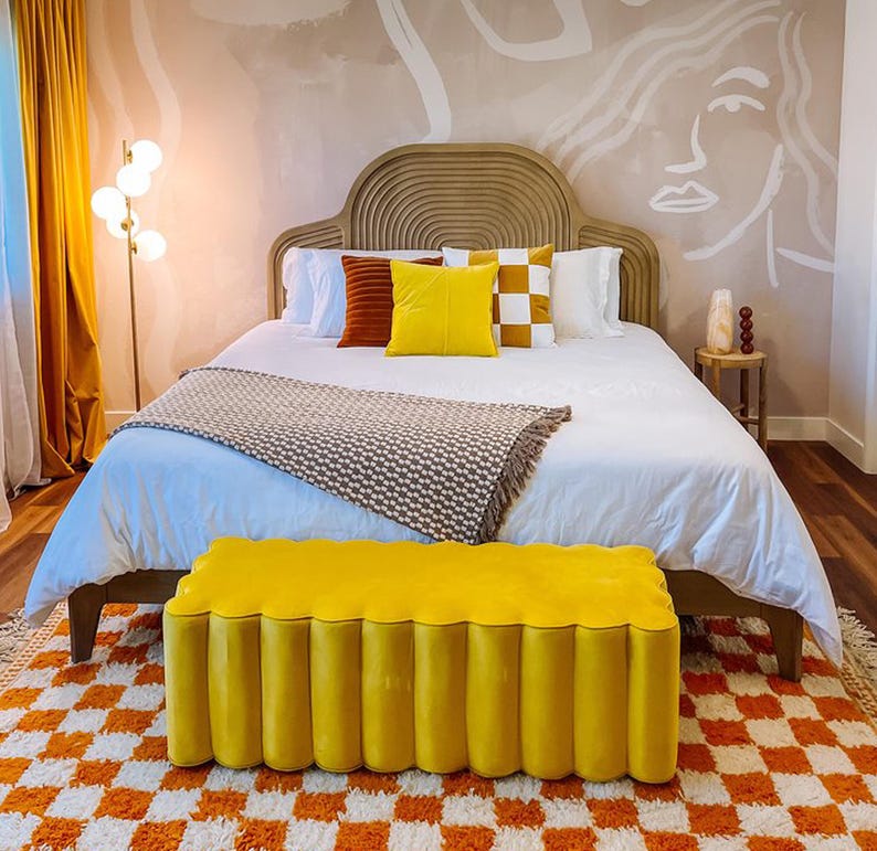 May include: A white bed with a brown headboard and a yellow tufted bench in front of it. The bed has a white blanket and a brown throw blanket. There are pillows on the bed in shades of brown, yellow, and white. The wall behind the bed is painted a light brown color and has a white abstract design on it.