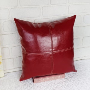 May include: A red leather pillow with white stitching. The pillow is square and has a smooth, shiny surface.
