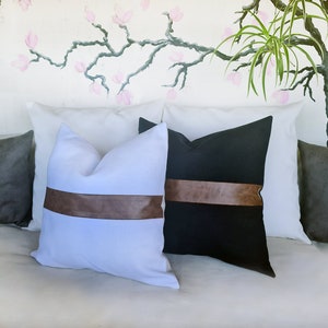 Puede incluir: Dos almohadas decorativas con ribete de cuero marr&oacute;n. Una almohada es blanca con una banda de cuero marr&oacute;n, y la otra es negra con una banda de cuero marr&oacute;n.