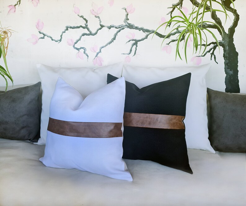 Puede incluir: Dos almohadas decorativas con ribete de cuero marr&oacute;n. Una almohada es blanca con una banda de cuero marr&oacute;n, y la otra es negra con una banda de cuero marr&oacute;n.