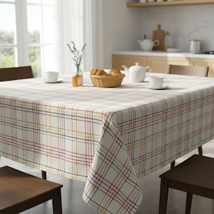 May include: A square tablecloth with a plaid pattern in cream, red, yellow, and blue covers a wooden table. A basket of croissants, a teapot, and two teacups are on the table. A vase of lavender and a jar of jam are also present.