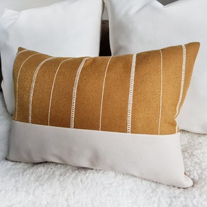 May include: A mustard yellow throw pillow with white stripes and a white faux leather bottom. The pillow is sitting on a white fluffy surface.