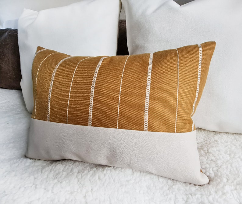 May include: A mustard yellow throw pillow with white stripes and a white faux leather bottom. The pillow is sitting on a white fluffy surface.