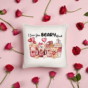 May include: White square pillow with the phrase "I Love You BEARY Much" and a graphic of teddy bears and coffee cups. The design includes hearts and the word "Love". Red roses are scattered around the pillow on a pink background.