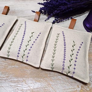 May include: Three small, white fabric pouches with lavender-themed embroidery. The pouches have brown leather ties and are filled with dried lavender.