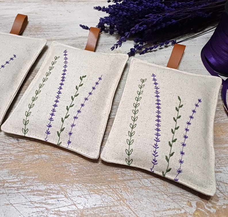 May include: Three small, white fabric pouches with lavender-themed embroidery. The pouches have brown leather ties and are filled with dried lavender.