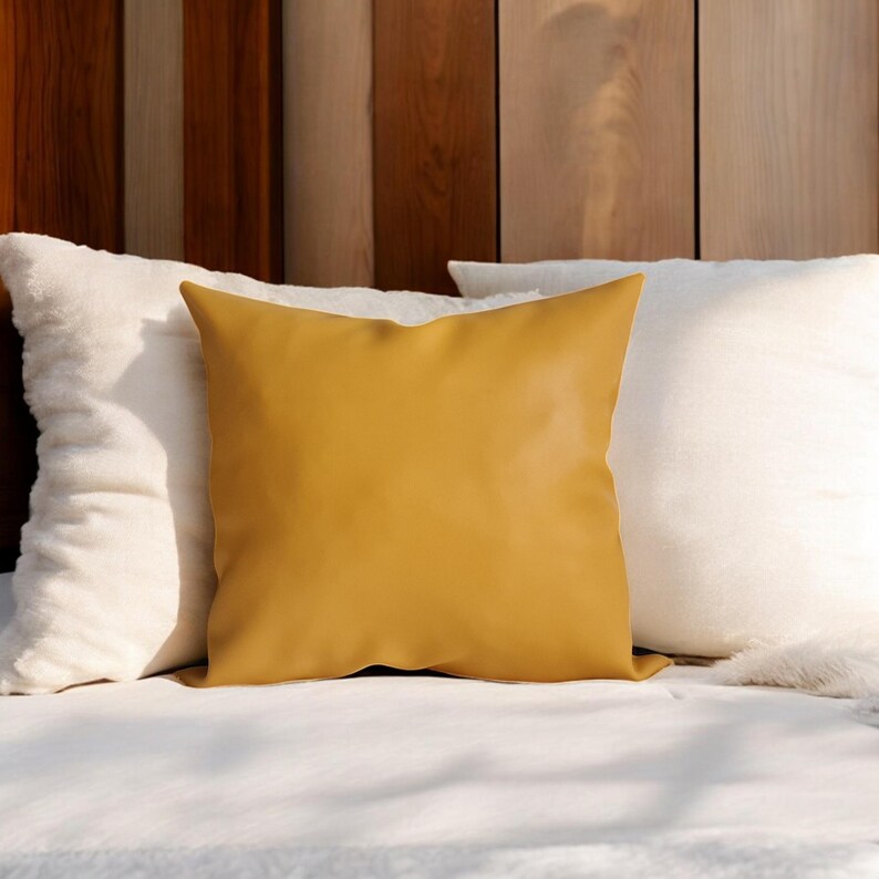May include: A mustard yellow throw pillow with a smooth, faux leather texture. The pillow is placed on a white bed with two white throw pillows behind it.