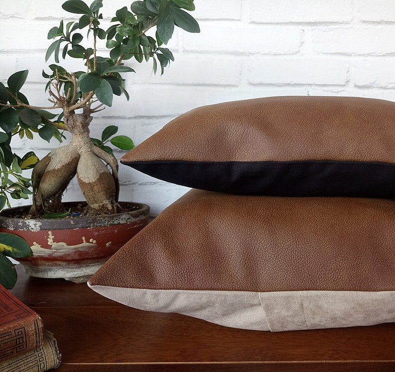 May include: Two brown faux leather throw pillows with black and beige trim. The pillows are stacked on top of each other.