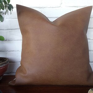 May include: A brown faux leather pillow with a textured surface. The pillow is square and has a simple design.