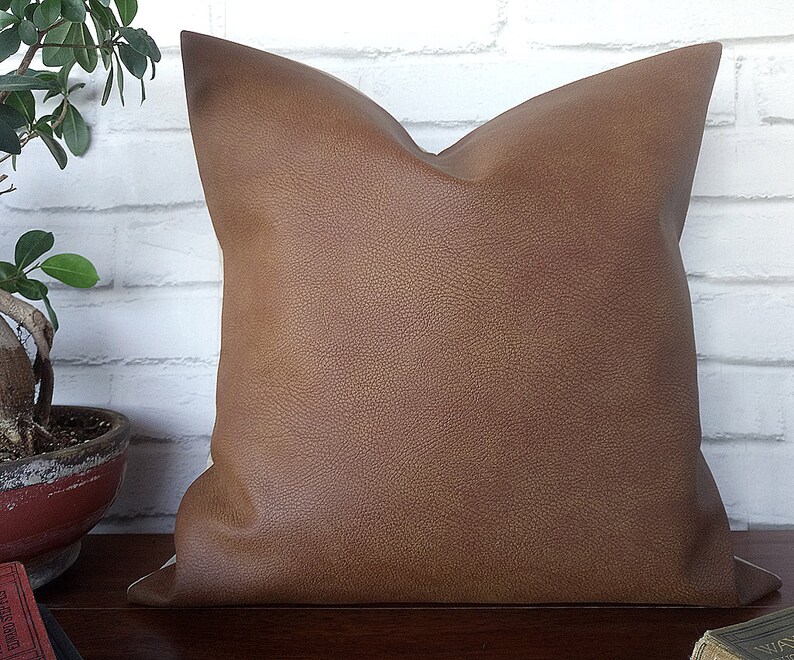 May include: A brown faux leather pillow with a textured surface. The pillow is square and has a simple design.