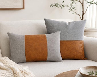May include: Two decorative throw pillows on a white sofa. One rectangular pillow features a brown leather center panel with gray fabric on the sides. The other pillow is gray on top and brown leather on the bottom.