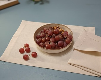 May include: A round ceramic bowl with a light-colored interior and a darker rim, filled with red grapes and walnuts. Four loose red grapes sit on a rectangular off-white linen placemat next to a folded matching linen napkin.
