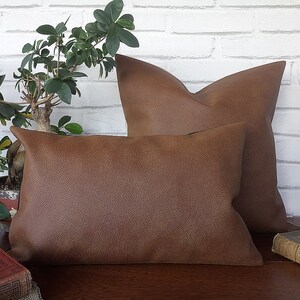 May include: Two brown faux leather throw pillows, one larger than the other, are arranged on a wooden surface. The pillows have a textured surface and are in focus.