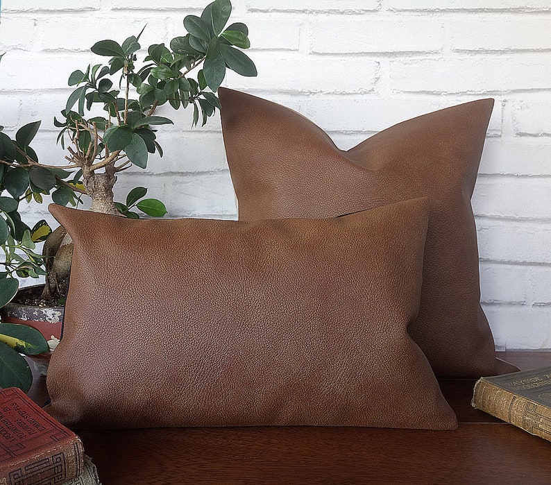May include: Two brown faux leather throw pillows, one larger than the other, are arranged on a wooden surface. The pillows have a textured surface and are in focus.