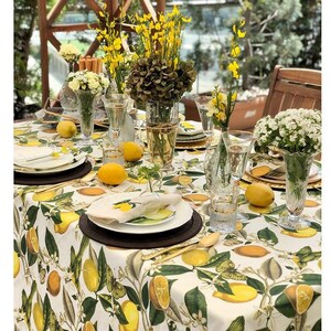 May include: A beautifully set table with a lemon-themed tablecloth, featuring lemons and green leaves. The table is adorned with white plates, gold cutlery, clear glasses, and vases of yellow and white flowers. Fresh lemons are scattered across the table.