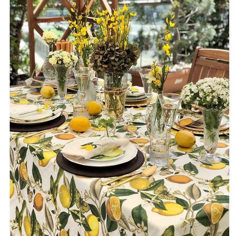 May include: A beautifully set table with a lemon-themed tablecloth, featuring lemons and green leaves. The table is adorned with white plates, gold cutlery, clear glasses, and vases of yellow and white flowers. Fresh lemons are scattered across the table.