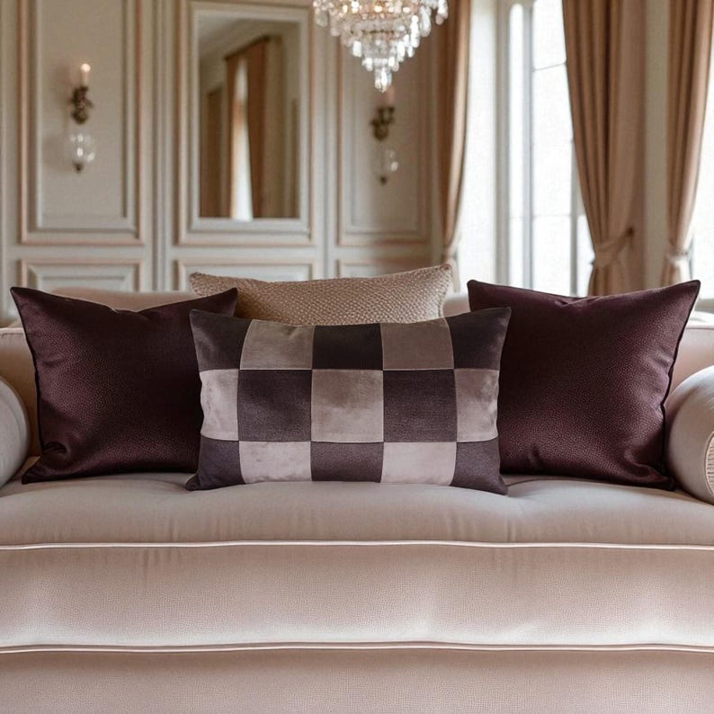 May include: A beige sofa with several decorative pillows. Two dark burgundy pillows with textured surfaces flank a checkered pillow in shades of brown and beige. A smaller, textured beige pillow sits in the background.