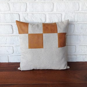 May include: A beige pillow with brown leather squares sewn on the front. The pillow is sitting on a wooden surface.