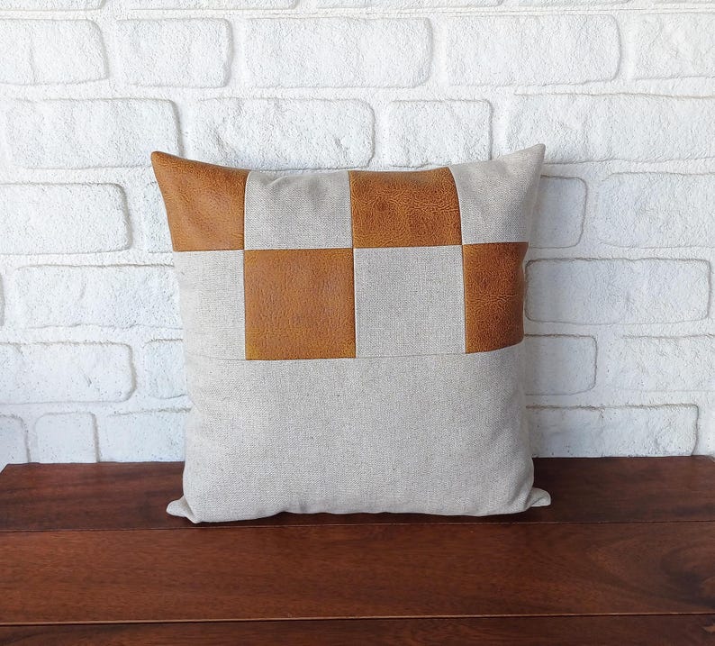 May include: A beige pillow with brown leather squares sewn on the front. The pillow is sitting on a wooden surface.