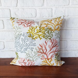 Pu&ograve; includere: Cuscino quadrato decorativo con un motivo a barriera corallina ricamato su sfondo bianco. Il corallo &egrave; nelle tonalit&agrave; di rosso, giallo, verde e nero. Il cuscino &egrave; appoggiato su una superficie di legno davanti a un muro di mattoni bianchi.