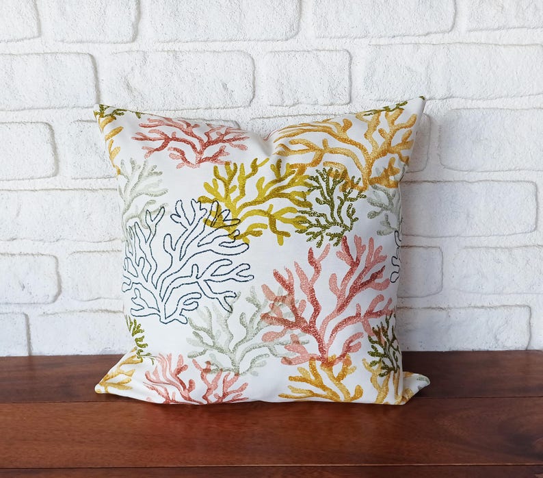 Pu&ograve; includere: Cuscino quadrato decorativo con un motivo a barriera corallina ricamato su sfondo bianco. Il corallo &egrave; nelle tonalit&agrave; di rosso, giallo, verde e nero. Il cuscino &egrave; appoggiato su una superficie di legno davanti a un muro di mattoni bianchi.