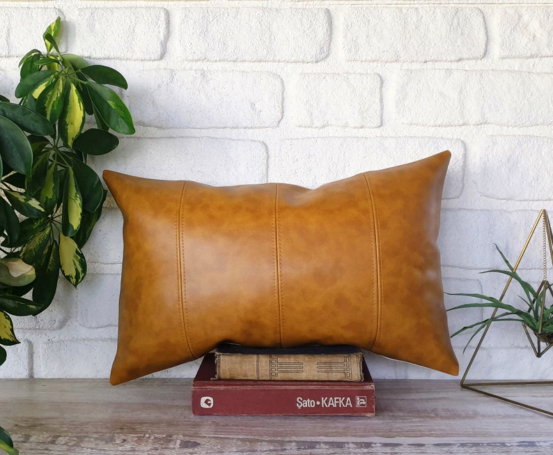 Puede incluir: Una almohada de cuero marr&oacute;n con forma rectangular y costuras. La almohada est&aacute; apoyada en una pila de libros.