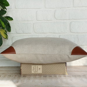 May include: A light brown linen pillow with brown leather accents on the corners. The pillow is sitting on top of a book with the text "Complete Sherlock Holmes" on the spine.