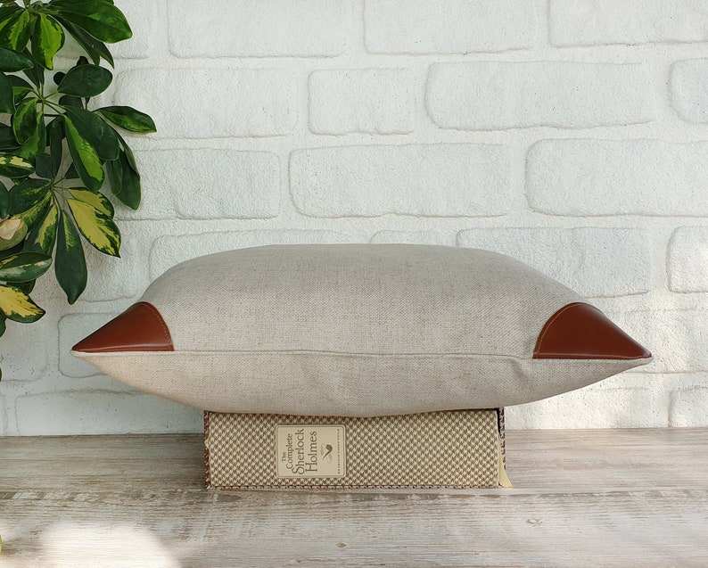 May include: A light brown linen pillow with brown leather accents on the corners. The pillow is sitting on top of a book with the text "Complete Sherlock Holmes" on the spine.