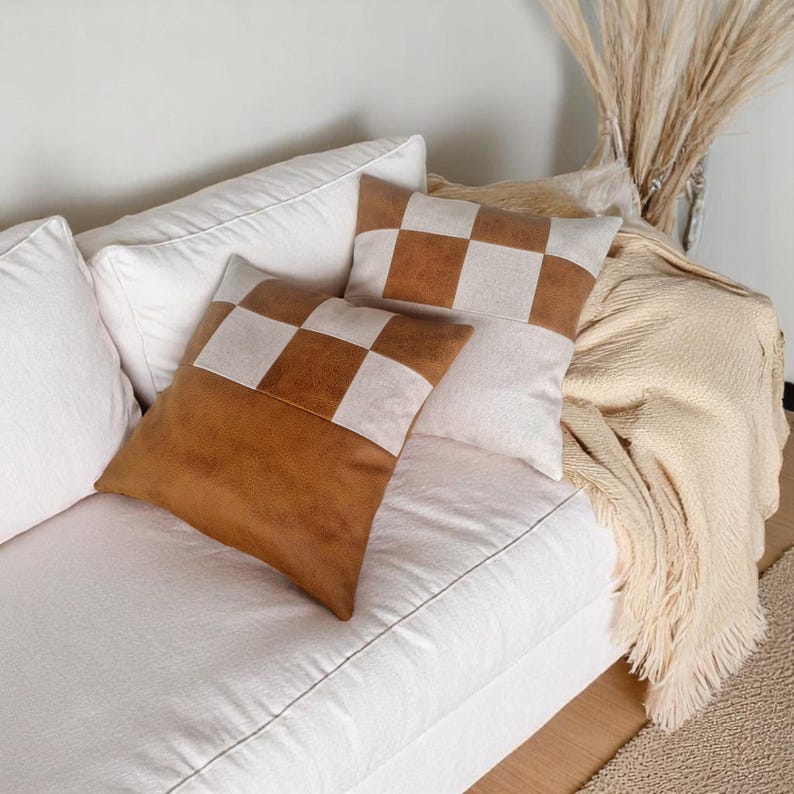 May include: Two brown and white checkered throw pillows with a suede-like texture are placed on a white couch. A cream-colored throw blanket with fringe is draped over the couch.