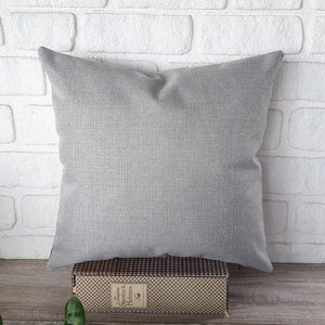 May include: A gray square pillow with a textured fabric surface. The pillow is sitting on a brown and white checkered box.