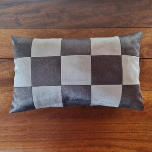 May include: A rectangular decorative pillow with a checkerboard pattern. The pillow features alternating squares of dark gray and light gray fabric. The pillow is placed on a wooden surface.