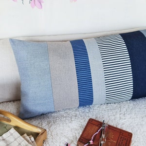 May include: A long rectangular pillow with a patchwork design. The pillow is made of different shades of blue denim and striped fabric. The pillow is lying on a white fluffy surface.