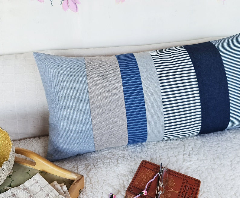 May include: A long rectangular pillow with a patchwork design. The pillow is made of different shades of blue denim and striped fabric. The pillow is lying on a white fluffy surface.