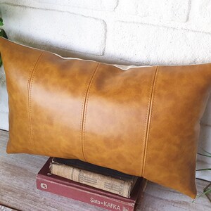 Puede incluir: Una almohada lumbar de cuero marr&oacute;n con un acabado texturizado sutil. La almohada tiene una forma rectangular con una costura en el centro y dos costuras a cada lado.
