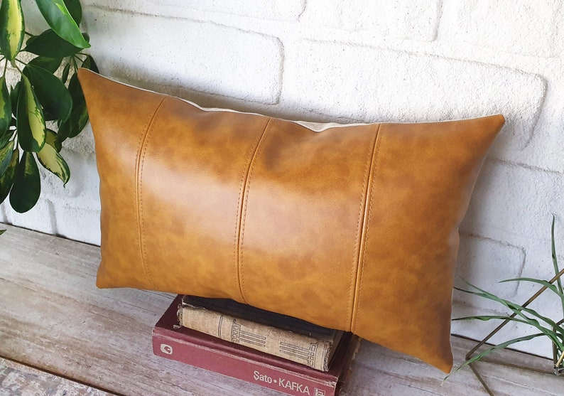 Puede incluir: Una almohada lumbar de cuero marr&oacute;n con un acabado texturizado sutil. La almohada tiene una forma rectangular con una costura en el centro y dos costuras a cada lado.