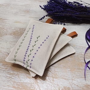 May include: Three beige linen sachets with lavender embroidery. The sachets have brown leather ties and are stacked on top of each other.