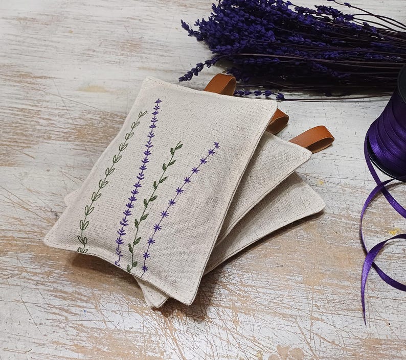 May include: Three beige linen sachets with lavender embroidery. The sachets have brown leather ties and are stacked on top of each other.
