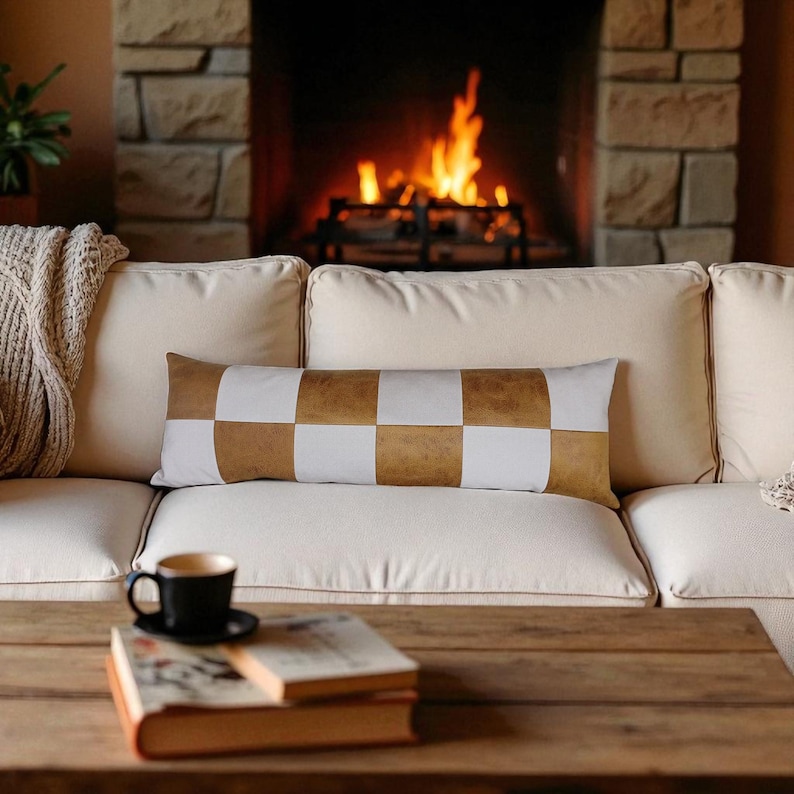 May include: A rectangular decorative pillow with a checkerboard pattern of white and tan squares rests on a cream-colored sofa. A burning fireplace is in the background. A black coffee cup and books are on a wooden coffee table.
