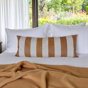 May include: A white bed with a brown and white striped lumbar pillow and a brown blanket. The bed is in front of a window with white curtains.