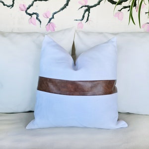Puede incluir: Almohada blanca con una banda de cuero marr&oacute;n en el centro. La almohada est&aacute; en un sof&aacute; blanco.