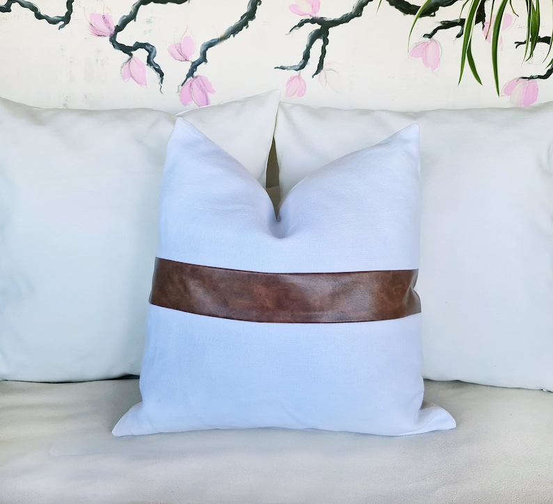 Puede incluir: Almohada blanca con una banda de cuero marr&oacute;n en el centro. La almohada est&aacute; en un sof&aacute; blanco.